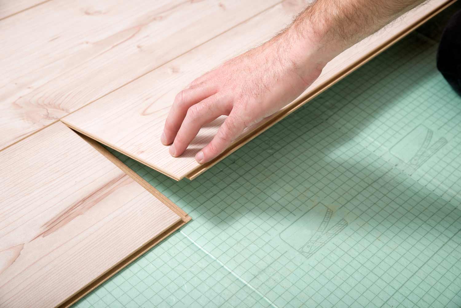 Hand legt eine helle Laminatdiele auf einer grünen Trittschalldämmung ein und klickt sie in das benachbarte Paneel ein.